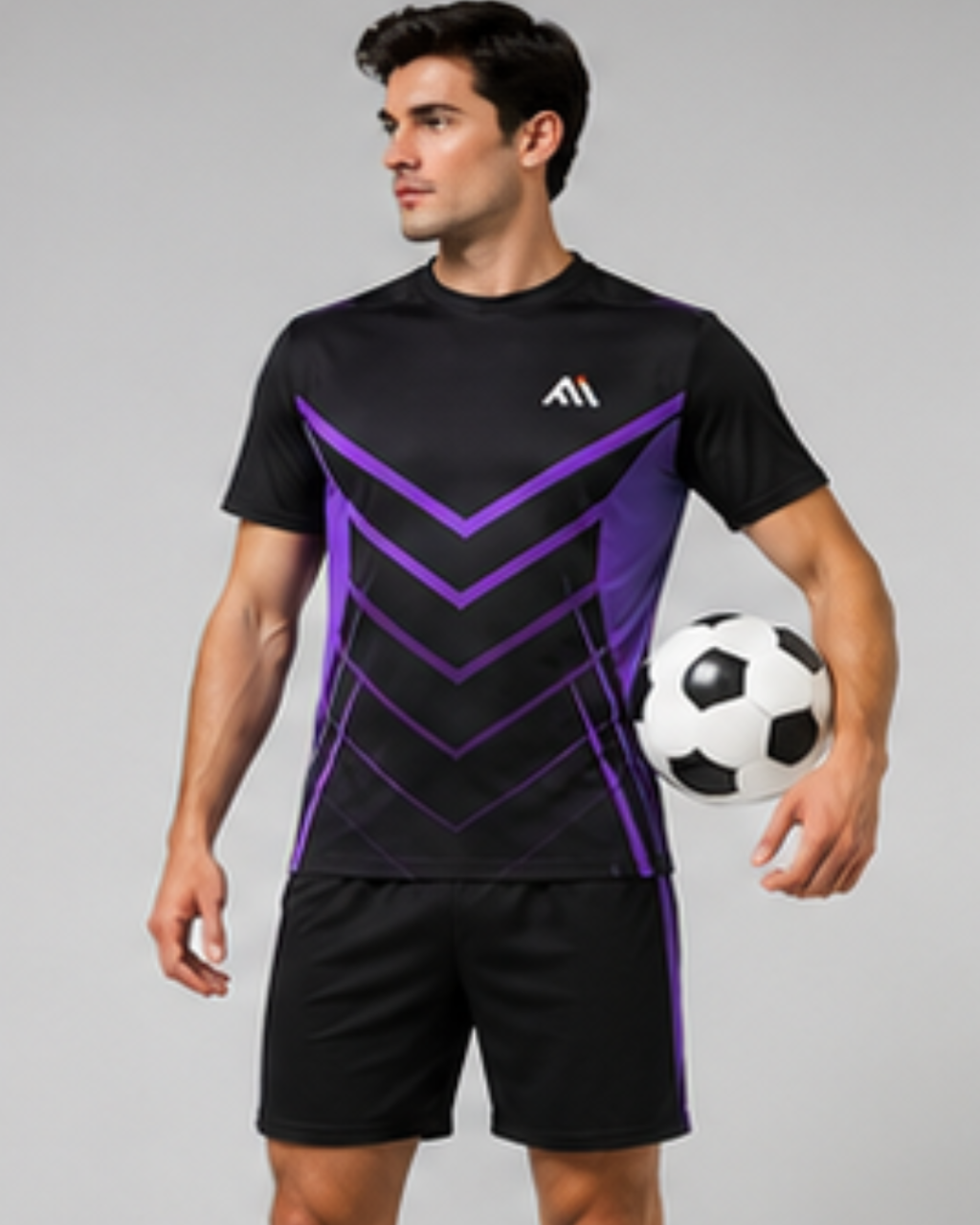 A man modeling a black athletic soccer jersey with purple chevron patterns and matching black shorts, holding a soccer ball against a plain gray studio background.