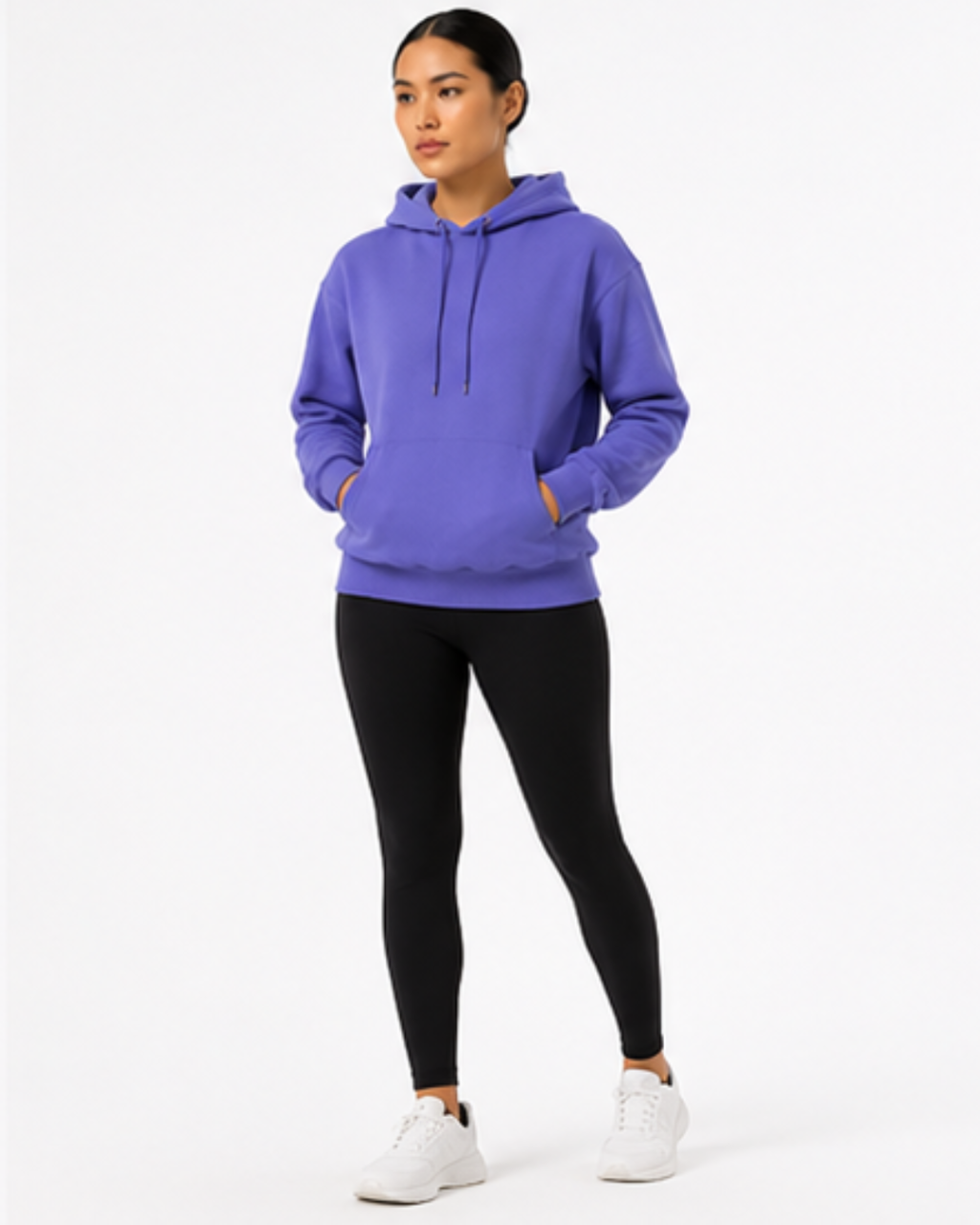 A woman modeling a purple pullover hoodie with black leggings and white sneakers, standing against a plain white studio background.